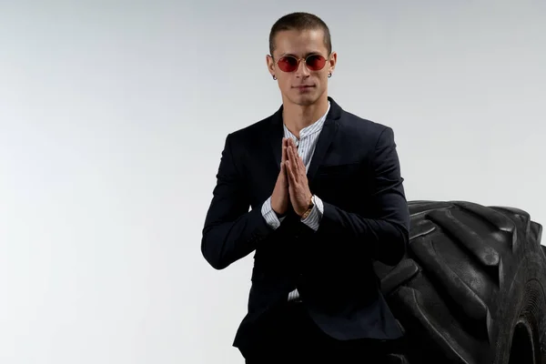 Hombre joven guapo en traje negro y gafas de sol rojas de pie delante de un neumático grande, mirando a la cámara y sosteniendo las manos juntas, aislado sobre fondo blanco, espacio de copia para su texto — Foto de Stock