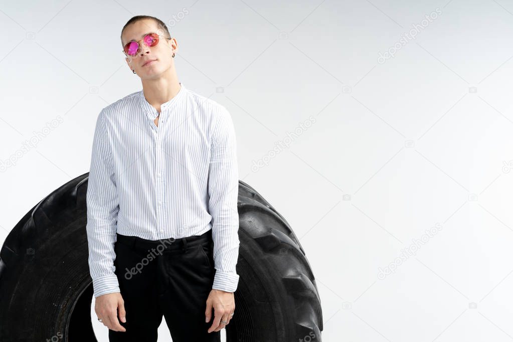 Handsome young man in white shirt in red sunglasses standing in front of the big tire, looking at the camera, isolated over white background, copyspace for your text