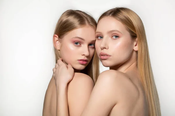 Dos mujeres gemelas atractivas sexy con el pelo largo y rubio posando en maquillaje glamour, de pie uno detrás del otro, posando en el estudio sobre fondo gris. Lesbianas sexy pareja — Foto de Stock