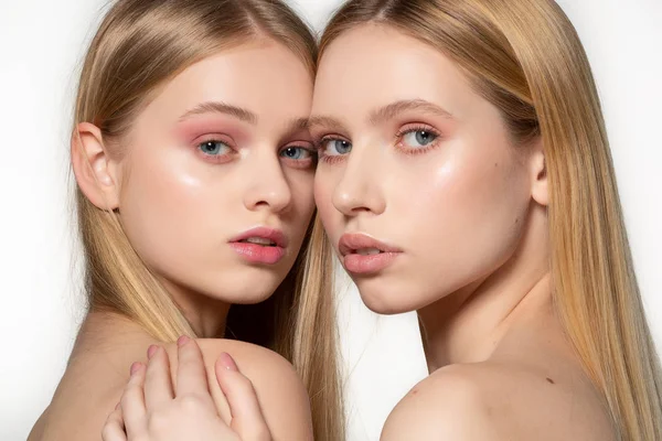 Dos mujeres gemelas atractivas sexy con el pelo largo y rubio posando en maquillaje glamour, de pie uno detrás del otro, posando en el estudio sobre fondo gris. Lesbianas sexy pareja — Foto de Stock