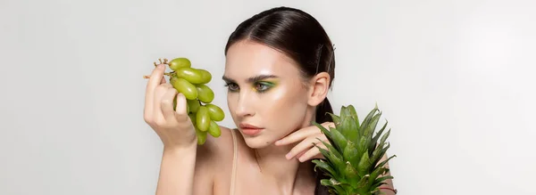 Friska unga brunett kvinna tittar på gröna druvor i hennes hand, frukt och grönsaker på bordet, studio foto på grå bakgrund — Stockfoto