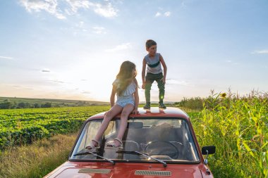 Kırmızı retro arabanın çatısında duran gri tişörtlü genç çocuk ve sol tarafta korkutucu görünümlü mavi çizgili elbise li kız kardeşi yanında oturan ve ona bakarak, alan ve mavi gökyüzü