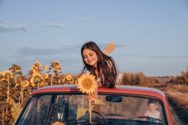 Çizgili mavi elbise güzel sevimli genç kız kırmızı retro arabanın çatısında giyinmiş gözleri ile yatıyor ve elinde ayçiçeği tutan, arka planda mavi gökyüzü