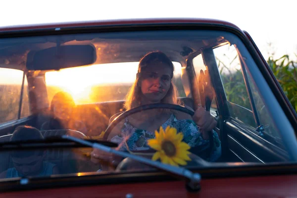 Beautiful blonde young mom driving a retro car with her happy children that enjoying summer travel, sunset on the bckground
