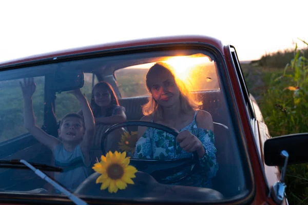 Linda loira jovem mãe dirigindo um carro retro com seus filhos felizes que gostam de viagens de verão, pôr do sol no chão — Fotografia de Stock