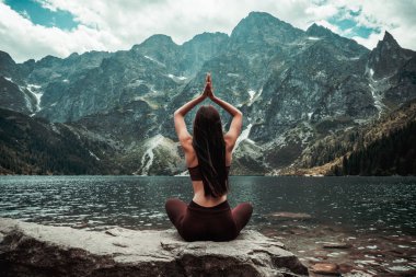 Koyu kırmızı spor giyim genç kadın doğada yoga pratik, kamera ve meditasyon, dağ gölü ve arka plan, aktif yaşam kavramı üzerinde mesafe geri taş üzerinde oturan