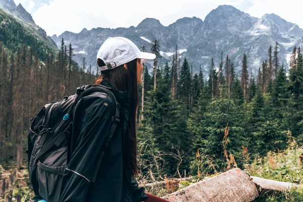 Bir sırt çantası ile beyaz şapkalı genç kız gezgin, güzel panorama zevk, dağlara gitmeden önce dinlenme, bir konkret çit üzerinde oturur — Stok fotoğraf