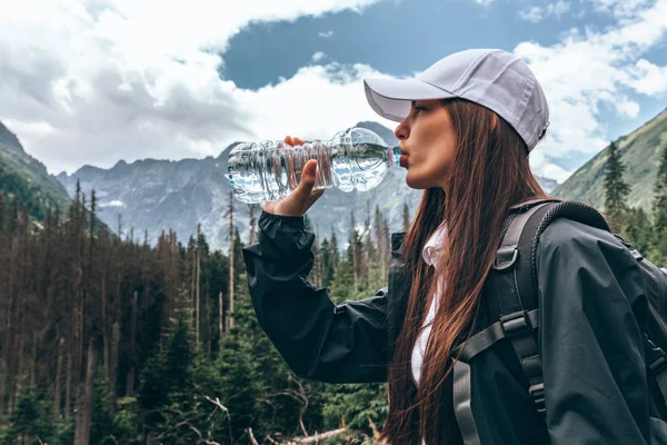 Beyaz şapka lı ve siyah ceketli genç güzel kız gezginin yan görünüm portresi içme suyu, arka planda güzel dağlar — Stok fotoğraf