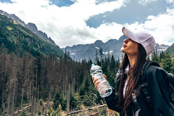 Beyaz şapka ve siyah ceket ve sırt çantası genç güzel kız gezgin, kapalı gözleri ile ayakta ve su bir şişe tutan, arka planda inanılmaz dağlar — Stok fotoğraf