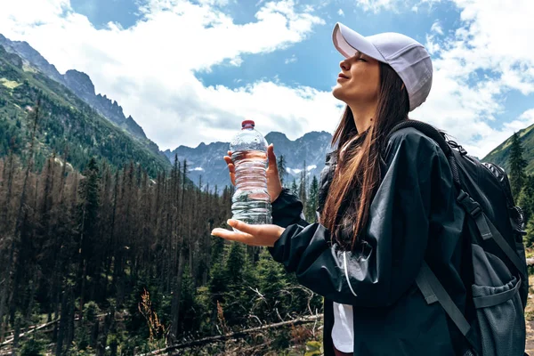 Beyaz şapka ve siyah ceket ve sırt çantası genç güzel kız gezgin, kapalı gözleri ile ayakta ve su bir şişe tutan, arka planda inanılmaz dağlar — Stok fotoğraf