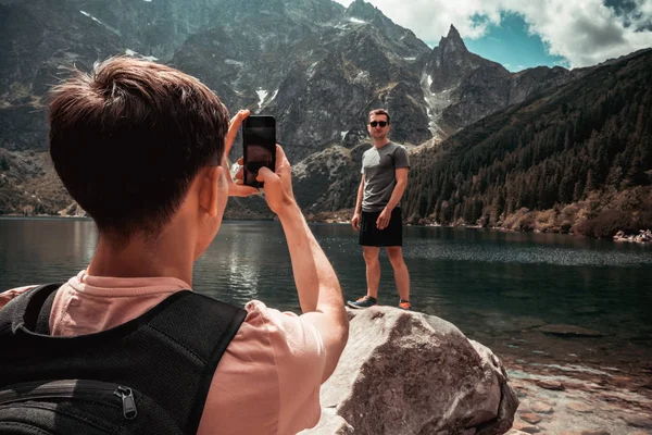 Two friends travelers in casual clothes, one man takes a photo of his friend that standing on the stone in front of the mountain lake