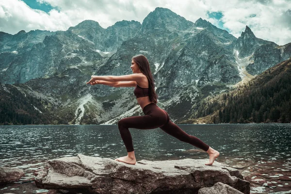 요가를 연습하는 젊은 매력적인 여성, 전사 한 운동에 서, Virabhadrasana 나는 포즈, 운동, 어두운 빨간 스포츠웨어를 입고, 산악 호수와 배경에 범위 — 스톡 사진