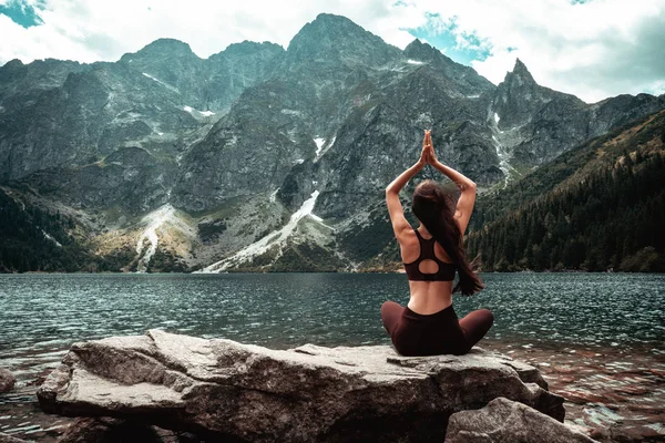 Koyu kırmızı spor giyim genç kadın doğada yoga pratik, kamera ve meditasyon, dağ gölü ve arka plan, aktif yaşam kavramı üzerinde mesafe geri taş üzerinde oturan — Stok fotoğraf