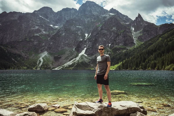 Siyah güneş gözlüğü ve gündelik giysiler dağ gölü ve aralığı önünde büyük taş üzerinde duran genç adam — Stok fotoğraf
