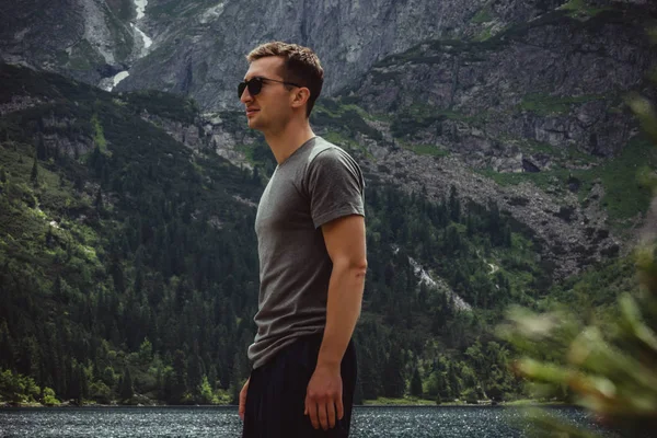 Retrato de vista lateral del joven viajero en gafas de sol negras y camiseta gris frente al lago de montaña y la cordillera, concepto de vida activa — Foto de Stock