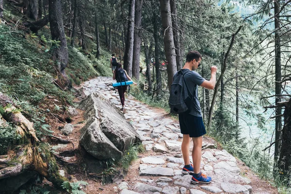 숲 길에 서있는 캐주얼 한 옷을 입은 젊은 남자 여행자는 아름다운 산악 호수의 전화 사진을 만듭니다. — 스톡 사진