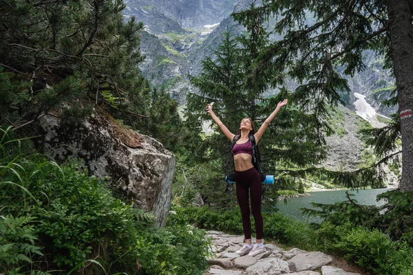 Όμορφο νεαρό σπορ κορίτσι ταξιδιώτη στέκεται με ανυψά τα χέρια στο μονοπάτι του δάσους μεταξύ των βουνών, λίμνη στο παρασκήνιο, ενεργό έννοια της ζωής — Φωτογραφία Αρχείου