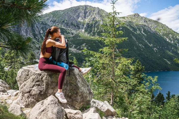 빨간 스포츠웨어에 젊은 매력적인 소녀는 큰 돌에 앉아, 그녀의 배낭에 기대어, 산을 등반하는 동안 피곤, 배경에 아름다운 산 호수, 활동적인 라이프 스타일 개념 — 스톡 사진