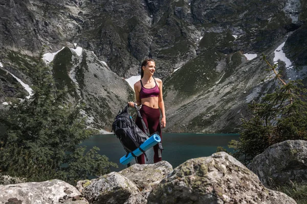 Młoda Dziewczyna Brunetka samotnie w czerwonym Sportswear z plecakiem w ręku stoi na kamieniach, niesamowite jezioro Mountine na tle, aktywny i zdrowy styl życia koncepcji — Zdjęcie stockowe