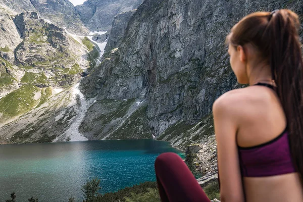 Zbliżenie Fotografia ofyoung atrakcyjna dziewczyna samotnie w czerwonej odzieży sportowej, która siedzi z powrotem do aparatu i patrząc na niesamowite górskie jezioro — Zdjęcie stockowe