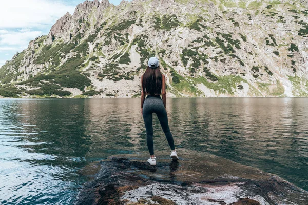 Güzel dağ gölü, aktif lifetyle kavramı yakınındaki büyük kaya üzerinde kameraya geri duran gri pantolonlu genç kadın gezgin — Stok fotoğraf