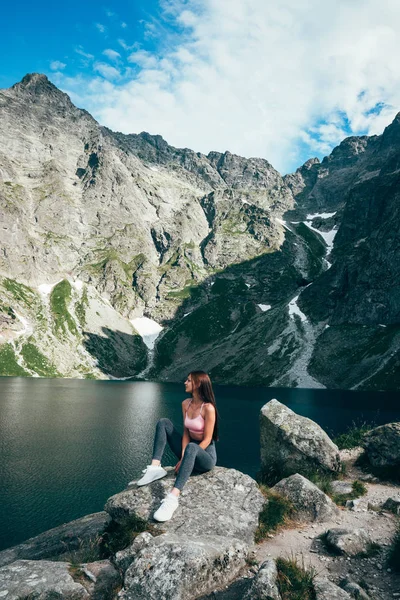 Gri pantolon genç güzel sportif kız büyük kaya üzerinde oturan ve dağ gölünün arka planda poz, aktif yaşam tarzı kavramı — Stok fotoğraf