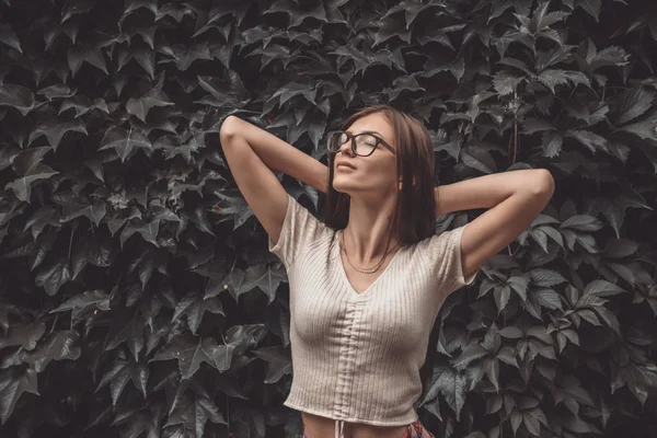 Half length portrait of beautiful young brunette girl in white t-shirt in glasses that holding hands behind head, leaf wall on the background
