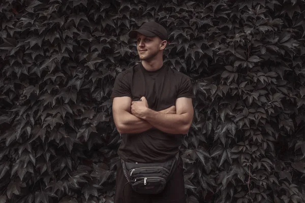 Hombre joven feliz en ropa casual y sombrero con bolso de hombro de pie con las manos cruzadas y mirando hacia otro lado, pared de la hoja en el fondo — Foto de Stock