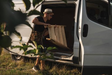 Omzunun üzerinde ulusal çanta ile beyaz şapka ve güneş gözlüğü genç adam çiftçi bir arabadan bir merdiven alır