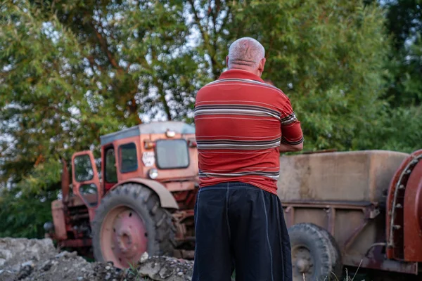 Ucraina, Chernivtsi, 10 08 2019 Uomo agricoltore o giardiniere in abiti casual in piedi dietro la macchina fotografica con le mani incrociate guardando il trattore rotto con spruzzatore serbatoio — Foto Stock
