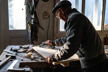 Gri giysi ve gözlük evde çalışan eski kıdemli marangoz, ahşap parlatır