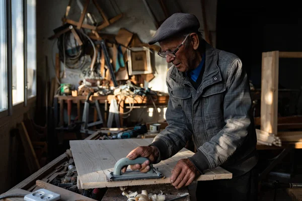 Muy viejo maestro carpintero en gafas pule madera en el taller del hogar, trabajando duro —  Fotos de Stock