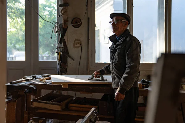 Porträt eines sehr alten Zimmermanns mit Brille in grauer Kleidung und Hut, der in der Nähe des Arbeitsplatzes mit Schleifwerkzeug in der Hand posiert — Stockfoto