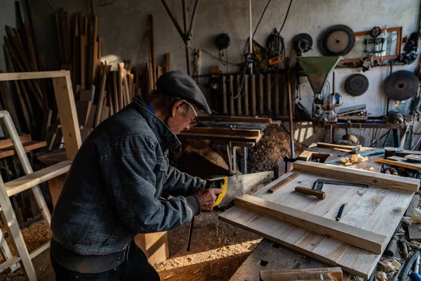 Molto vecchio maestro falegname in occhiali e abiti grigi che lavora duramente nella sua officina di casa, segheria — Foto Stock