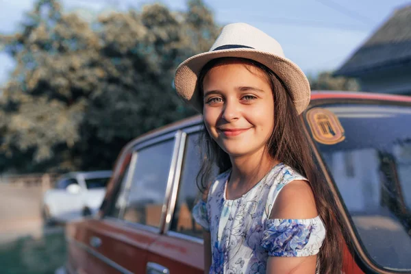 Portrait claudé, belle jeune fille brune en robe et chapeau appuyé sur la vieille voiture rouge et regardant la caamera — Photo