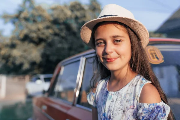 Portrait claudé, belle jeune fille brune en robe et chapeau appuyé sur la vieille voiture rouge et regardant la caamera — Photo