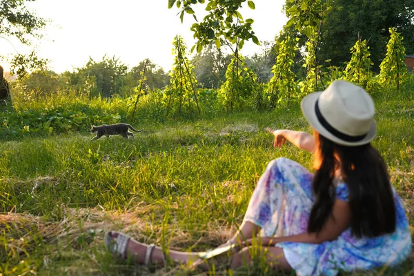 Jeune adolescente en robe avec imprimé de fleurs et chapeau blanc assis à l'arrière de la caméra sur la prairie avec herbe sèche tondue et pointant vers le chat gris — Photo