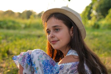 Beyaz şapka ve çiçek li elbise giyen sağlıklı bir genç kızın portresini kapatın parkta yerde oturan ve uzağa bakan baskılar