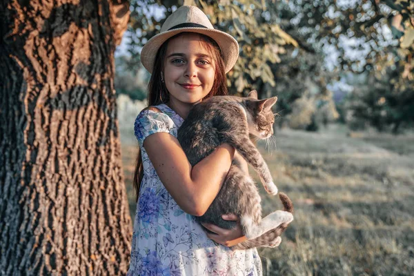 Felice sorridente giovane ragazza in cappello bianco e vestito con fiori di stampa che tiene un gatto bianco e grigio sulle mani, guardando la fotocamera, in piedi vicino al grande albero — Foto Stock