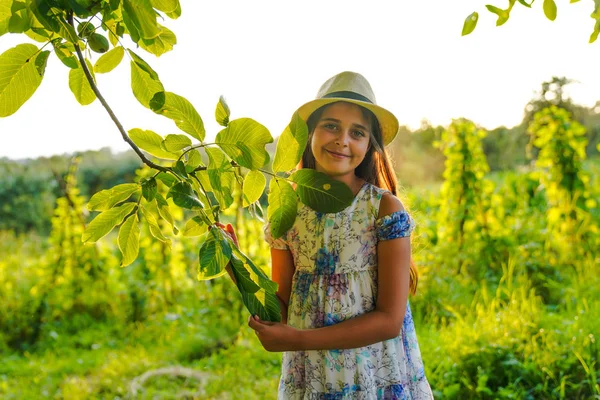 Feliz chica hermosa adolescente en sombrero blanco y vestido con flores imprimir la celebración de la rama, sonriendo y mirando a la cámara, pasar tiempo en el parque soleado —  Fotos de Stock