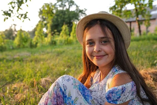 Joyeux sourire jeune fille aux cheveux bruns en chapeau blanc et robe avec des fleurs imprimé assis sur le sol dans le parc — Photo