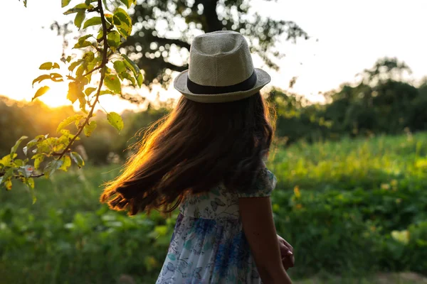 Teen girl in white hat and dress standing in the park back to the camera with loose hair and looking at sunset