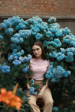 Genç güzel esmer kız pembe t-shirt ve bej pantolon mavi çiçekler abd arasında oturan kamera bakarak giymiş