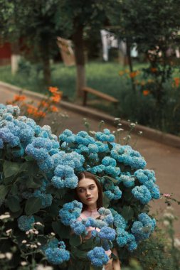 Mavi çiçekler arasında duran esmer saçlı neşeli genç kadın