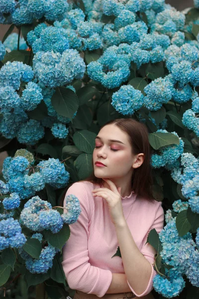 Seriamente bela menina morena com maquiagem brilhante em camiseta rosa entre flores azuis, tocando seu rosto com a mão — Fotografia de Stock