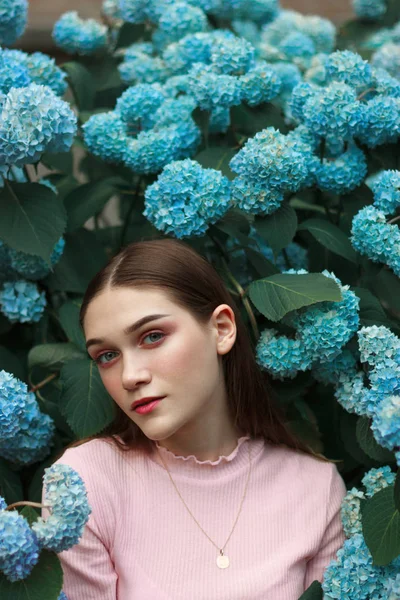 Primer plano retrato de la hermosa joven morena con maquillaje brillante usando camiseta rosa de pie entre flores azules y mirando a la cámara —  Fotos de Stock