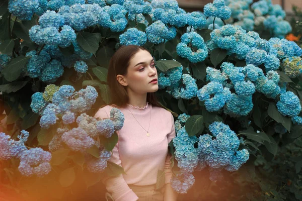 Zmysłowy, Młoda brunetka dziewczyna z jasnym makijażu noszenie ping t-shirt, stojący wśród niebieskich kwiatów i patrząc z dala — Zdjęcie stockowe