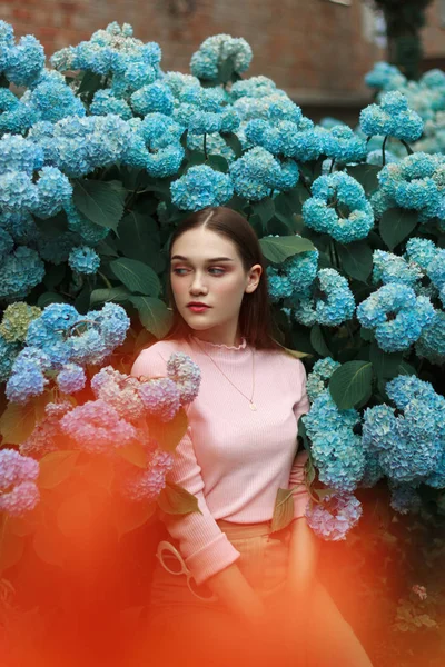 Sensual, joven morena con maquillaje brillante vistiendo camiseta de ping, de pie entre flores azules y mirando hacia otro lado —  Fotos de Stock