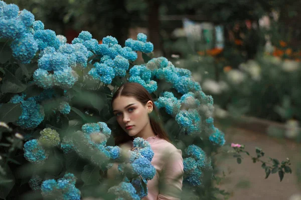 Belle fille brune aux lèvres rouges et aux yeux bleus debout parmi les fleurs bleues et regardant la caméra — Photo