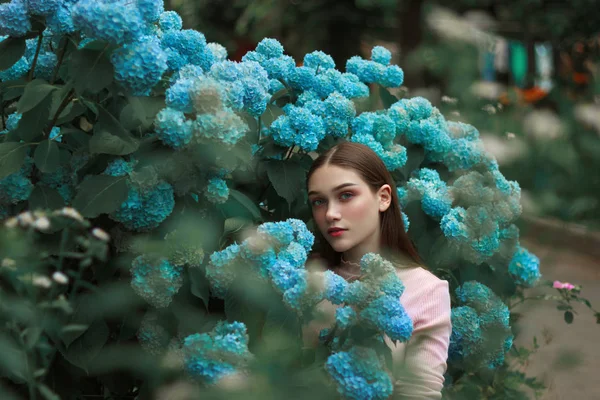 Belle fille brune aux lèvres rouges et aux yeux bleus debout parmi les fleurs bleues et regardant la caméra — Photo